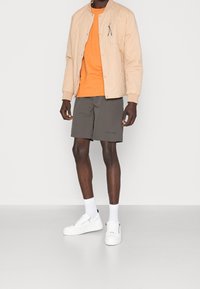Veste bomber matelassée beige, t-shirt orange, shorts gris et sneakers blancs avec semelles texturées ; le mannequin se tient devant un fond blanc.
