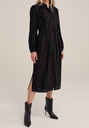 Vrouw draagt een zwart midi-jurk met lange mouwen, een ceintuur en een subtiel blauw patroon, gecombineerd met zwarte, puntige enkellaarzen met hoge hak.