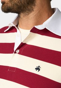 Pruhovaná polo košile v červené a krémové barvě s bílým límcem, knoflíkovou légou a malým vyšitým logem na hrudi. Materiál z bavlněné směsi.