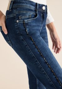 Jeans de mezclilla azul oscuro con un corte ajustado. Presentan costuras laterales con un patrón contrastante y el clásico diseño de cinco bolsillos.