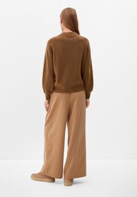 Femme se tenant de dos, portant un pull marron, un pantalon beige large et des baskets beiges, contre un fond blanc uni.
