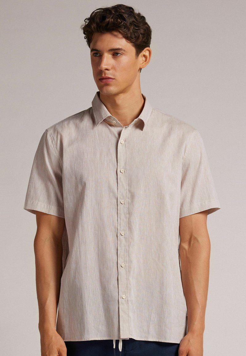 Camisa de botones de manga corta en beige claro con una textura sutil y rayas horizontales. Presenta un cuello clásico y seis botones en la parte delantera.