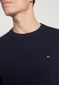 Pull en maille bleu marine avec un col rond et des poignets côtelés. Présente un petit patch logo aux couleurs rouge, blanc et bleu sur la poitrine.