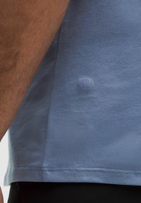 Tricou scurt cu mânecă, de culoare albastră, realizat dintr-un material neted, având o mică etichetă cu logo aproape de tiv și o croială lejeră, cu margini cusute.