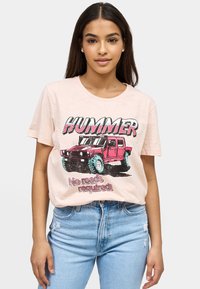 Lichtroze t-shirt met een grafische afbeelding van een rode Hummer met de tekst "HUMMER" en "Geen wegen nodig!" in vet, zwarte letters.