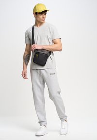 Herenoutfit met een lichtgrijze T-shirt en sweatpants, een gele pet en een zwarte schoudertas met een gele ritssluiting. Witte sneakers.