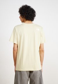 Kortærmet t-shirt i lyse beige bomuld, med et lille hvidt logo på den øverste ryg. Løs pasform og glat tekstur.