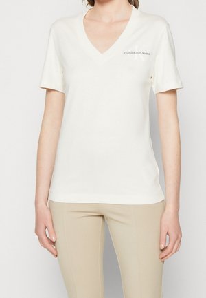 Femme portant un t-shirt blanc à manches courtes avec un col en V et un pantalon beige, le t-shirt comporte un petit logo Calvin Klein Jeans sur la poitrine.