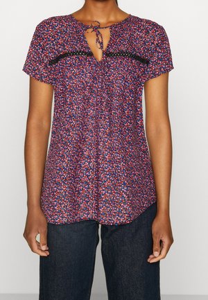 Vrouw draagt een korte mouwen blouse met een rood, blauw en roze bloemenpatroon en een klein strikje bij de halslijn, gecombineerd met een donkere spijkerbroek.