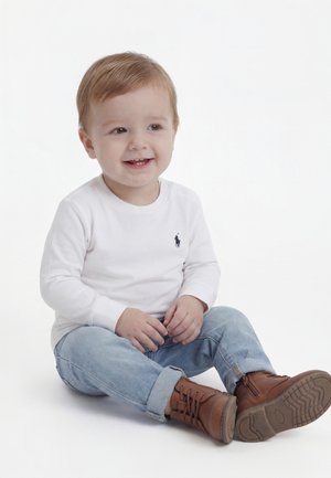 Malček sedi na tleh in se smehlja, oblečen v belo majico z dolgimi rokavi, svetlo modre kavbojke in rjave čevlje z vezalkami.