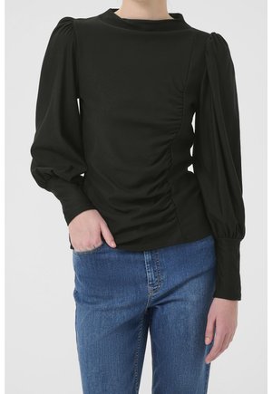 Woman wearing black ruched long-sleeve blouse with puffed shoulders and blue jeans, hand in pocket, against plain background.