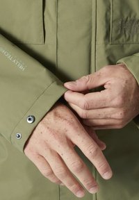 Groene waterdichte jas met een drukknoopmanchet, subtiele stikdetails en een logoopdruk op de mouw. Close-up van handen die de manchet aanpassen.