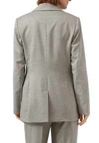 Vrouw in een lichtgrijze op maat gemaakte blazer en bijpassende broek, van achteren zichtbaar tegen een witte achtergrond.