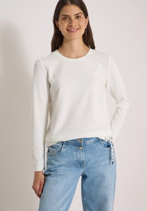 Femme aux longs cheveux foncés portant un haut blanc à manches longues et un jean bleu clair, debout avec une main dans la poche sur un fond uni.