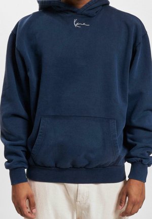 Sweat à capuche bleu marine avec poche avant et petit logo blanc sur la poitrine, porté avec un pantalon clair par une personne ayant les mains détendues le long du corps.