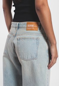 Helle blaue Denim-Jeans mit hoher Taille, fünf Taschen und einem Lederpatch auf der Rückseite mit Markenlogo und Text. Weicher Stoff, lässige Passform.