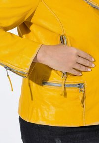 Chaqueta de cuero amarilla con cremalleras plateadas. Presenta un diseño ajustado, bolsillos con cremallera y una textura suave. Primer plano de una mano sobre la chaqueta.