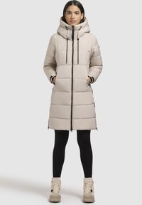 Heledbeez puffer-jakk, millel on kõrge krae ja nöörid, koos friteerimisega sektsioonide ja küljetaskutega, kombineeritud mustade retuuside ja heledate beezide saapadega.