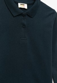 Tummansininen polo-paita, joka on valmistettu weikauspuuvillakankaasta. Siinä on kaulus, kolme napinpainiketta ja pieni tuotemerkki etiketti kaula-aukossa.