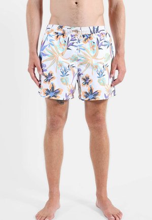 Hombre con pantalones cortos de baño blancos con patrones florales en naranja, azul y morado, de pie descalzo contra un fondo liso.