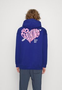 Modrá mikina s kapucí s velkým růžovo-bílým grafikou na zádech s nápisem "SO CHARMING" a srdcovým motivem, vyrobená z měkké látky.