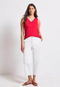 Czerwona bluzka bez rękawów z dekoltem w szpic, w zestawieniu z białymi, skróconymi spodniami. Brązowe płaskie sandały. Prosty, czysty design bez widocznych wzorów.