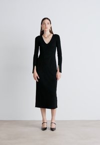 Black, fitted, long-sleeved dress with a V-neck, mid-length hem, textured fabric, paired with black heeled shoes and chunky gold jewelry.