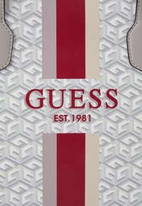 Väskan med logotryck har ett vitt geometriskt mönster och vertikala ränder i röd, beige och ljusgrå färg, med "GUESS EST. 1981" i fet röd text.