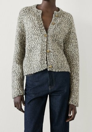 Personne portant un cardigan gris en grosse maille avec des boutons dorés, associé à un jean bleu foncé taille haute sur un fond uni.