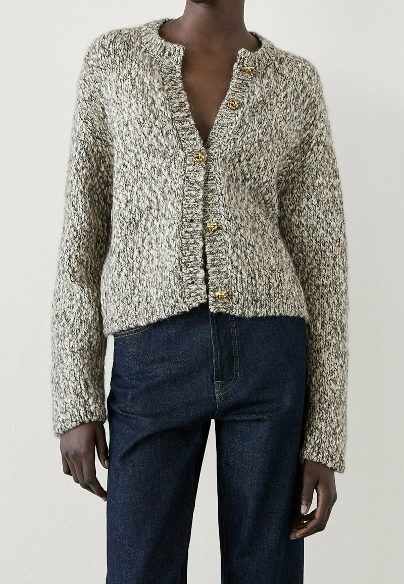 Personne portant un cardigan gris en grosse maille avec des boutons dorés, associé à un jean bleu foncé taille haute sur un fond uni.