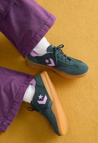 Baskets en daim vert avec des accents roses et un logo étoile, dotées d'une semelle en caoutchouc et de lacets noirs, associées à un pantalon violet et des chaussettes blanches.