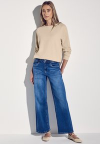 Maglia beige a maniche lunghe con polsini a coste, abbinata a jeans in denim blu a gamba larga con finitura sbiadita e orlo sfrangiato. Scarpe basse chiare.