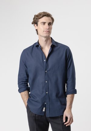 Hombre con cabello castaño claro que lleva una camisa azul oscuro abotonada con las mangas remangadas y pantalones negros, de pie frente a un fondo gris liso.