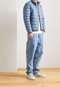 Giacca a vento blu con cuciture orizzontali, colletto grigio, t-shirt bianca, jeans azzurri chiari e sneakers grigie su un pavimento di legno.