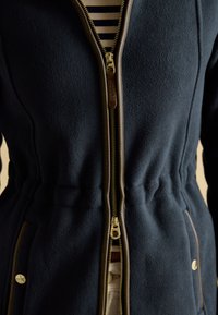 Primo piano di una giacca in pile blu navy con doppia cerniera bordata di marrone parzialmente aperta, che mostra una camicia a righe sotto.