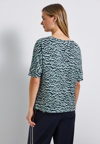 Nainen, jolla on pitkät vaaleat hiukset, pukeutuneena lyhythihaiseen vihreä-mustaan kuvioituun paitaansa ja tummiin housuihin, joissa on vaaleat raidat, katsoo hieman vasemmalle.