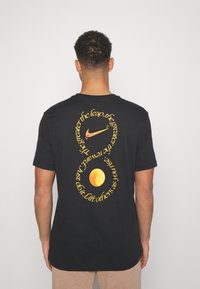 Svart bomull t-shirt med gul grafisk text och cirkulär design på baksidan, med en Nike-logotyp ovanför texten. Kort ärm.