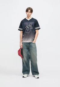 Ναυτικό και γκρι gradient αθλητικό φανελάκι με λευκό γιακ�ά και ρίγες. Χαλαρές τζιν παντελόνες και κόκκινο καπέλο κρατημένο στο πλάι. Sneakers συμπληρώνουν την εμφάνιση.