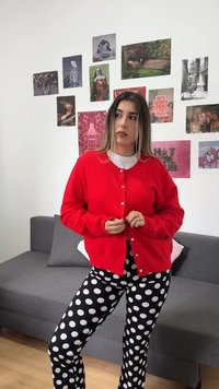 Νεαρή γυναίκα με κόκκινο ζακέτα και ασπρόμαυρο πουά παντελόνι στέκεται σε εσωτερικό χώρο κοντά σε γκρι καναπέ με καλλιτεχνικές φωτογραφίες σε λευκό τοίχο.
