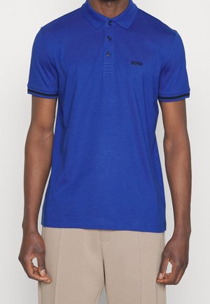 Homme portant un polo bleu cobalt à manches courtes avec une bordure noire sur les manches et le logo "BOSS" sur la poitrine, associé à un pantalon beige.