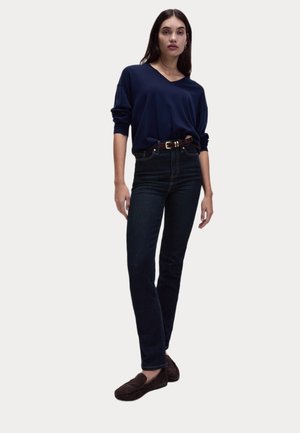 Pull à col en V marine, jean en denim foncé, ceinture marron et chaussons sombres. Tenue décontractée avec une coupe relaxée et une taille définie.
