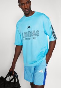 Šviesiai mėlynas tinklinis sportinis marškinėlis su juodu užrašu "ADIDAS SPORTSWEAR", derinamas su atitinkamais mėlynais šortais su baltais ir laivyno mėlynumo akcentais.