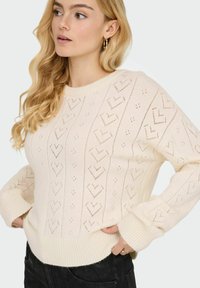 Pull en maille crème avec motifs en forme de cœur et poignets côtelés. Présente un col rond et un design texturé avec des détails ajourés.