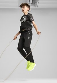 Čierny športový outfit so grafickým tričkom a nohavicami so sťahovacou šnúrkou, doplnený neonovo zelenými teniskami a švihadlom.