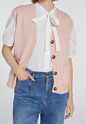 Woman wearing a white blouse with a tied bow neck, a pink buttoned vest, and high-waisted blue jeans, hand in pocket.
