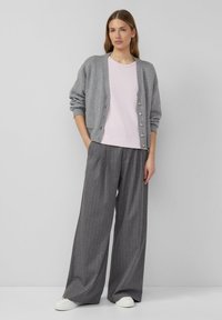 Graue Strickjacke mit Knöpfen, kombiniert mit einem hellrosa T-Shirt und grauen, gestreiften Hosen mit weitem Bein. Weiße Turnschuhe komplettieren das Outfit.