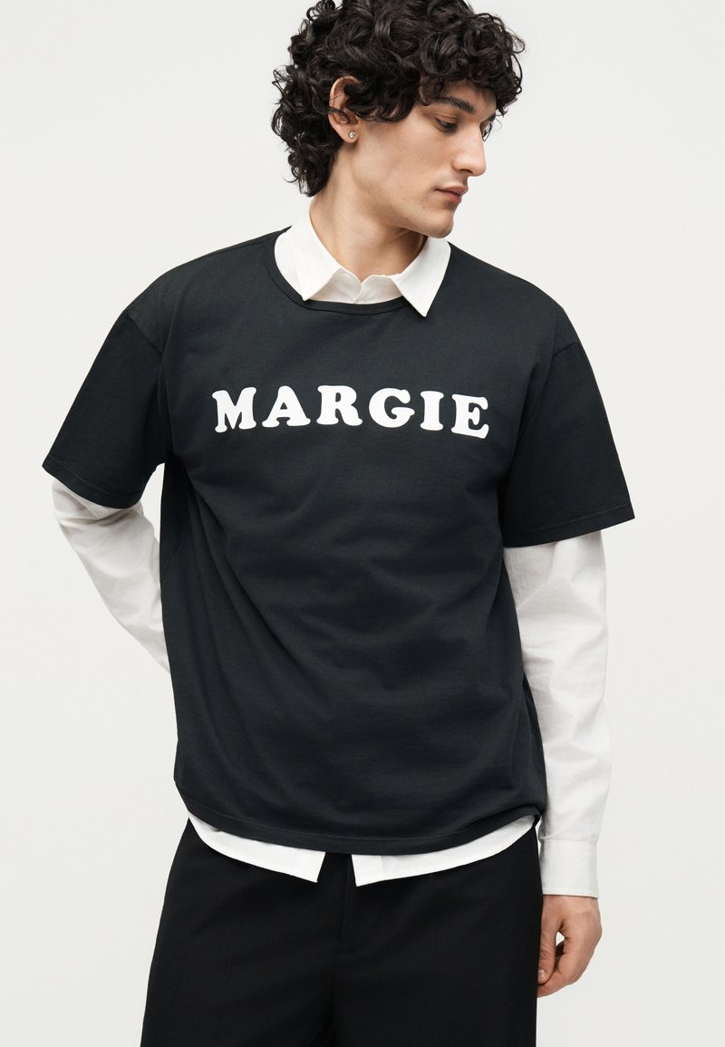 Jeune homme aux cheveux bouclés regarde vers la droite, portant un T-shirt noir avec « MARGIE » en blanc sur une chemise blanche à col et un pantalon noir.