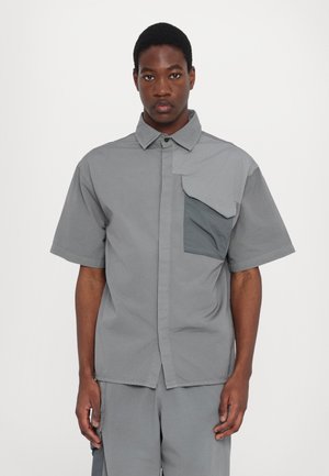 Short-sleeve, button-up shirt in light grey fabric with uneven patch pocket in darker grey, featuring a classic collar and relaxed fit.