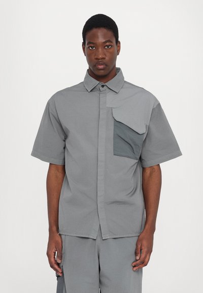 Camisa de manga corta, con botones, en tejido gris claro con un bolsillo de parche desigual en gris oscuro, que presenta un cuello clásico y un ajuste relajado.