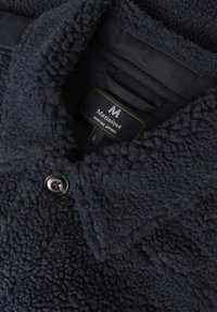 Tamsiai mėlynas pūkų švarkas su tekstūruota paviršiumi, su sagute ant apykaklės ir etiketė, skelbianti "Matinique Heritage Apparel, L."